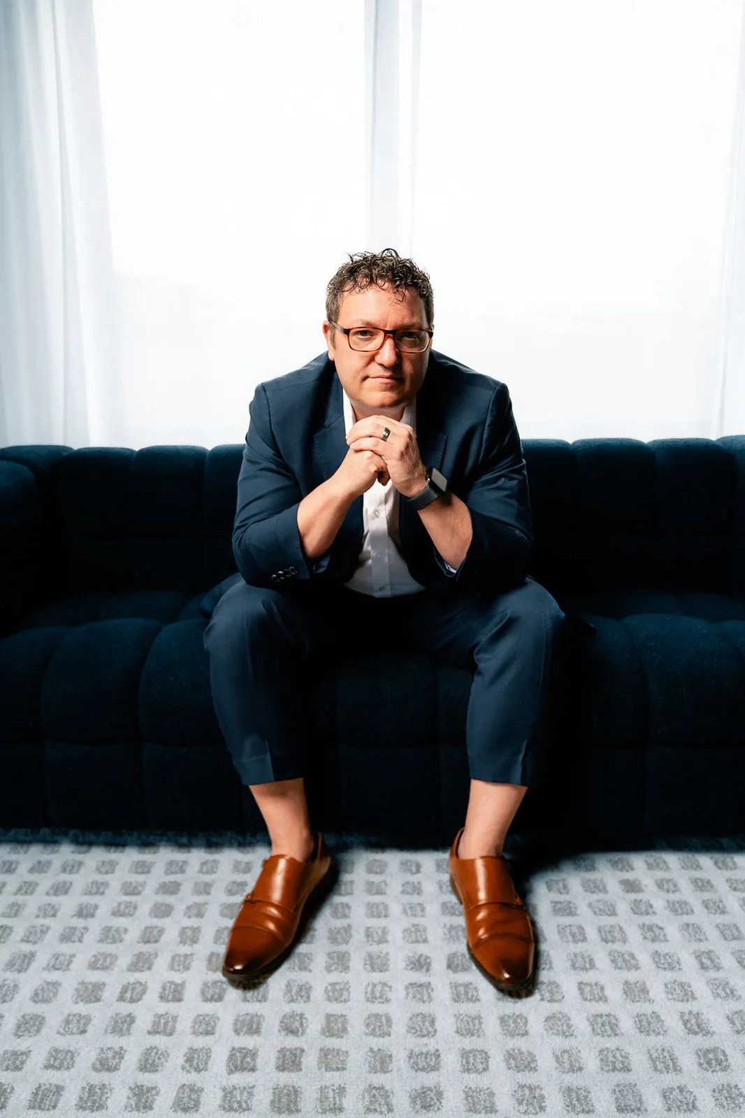 Billy Mawhiney seated on a dark sofa in a navy suit with his hands clasped